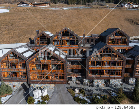 Large multi story Alpine hotel with gabled roofs, balconies, and a wooden facade, surrounded by snow, grassy hills, and rural buildings in the Dolomites. 129564688