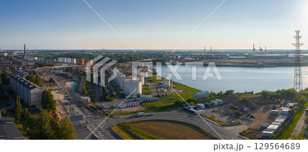 View of an industrial zone near a river in Riga, Latvia, featuring storage tanks, cranes, a shipyard, a curved road, and a tall transmission tower. 129564689