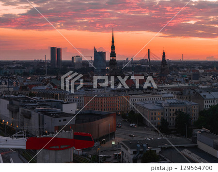 Riga, Latvia's old town skyline at sunset, featuring St. Peter's Church, Vansu Bridge over the Daugava River, and a mix of historic and modern buildings. 129564700