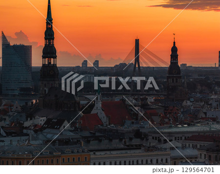 A sunset view of Riga, Latvia, featuring St. Peter's Church, Riga Cathedral, and the Vansu Bridge over the Daugava River, framed by diverse architecture. 129564701