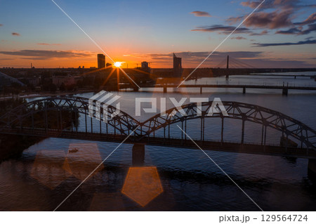 The image shows a sunset over Riga, Latvia, with the Railway Bridge spanning the Daugava River and the Vansu Bridge visible in the background. The image shows a sunset over Riga, Latvia, with the Railway Bridge spanning the Daugava River and the Vansu Bridge visible in the background. 129564724