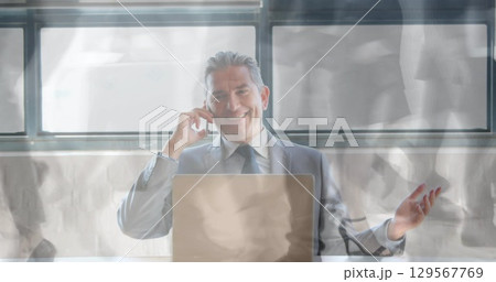 Smiling man in business suit holding smartphone to ear and gesturing at office desk, with laptop 129567769