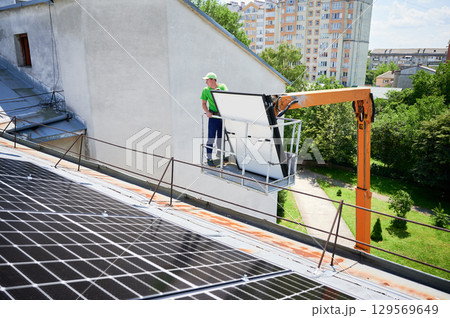 Workers lifting up photovoltaic solar panel on metal rooftop of house with assistance of crane lift. Men installers installing photovoltaic solar modules outdoors. Renewable energy generation concept. 129569649