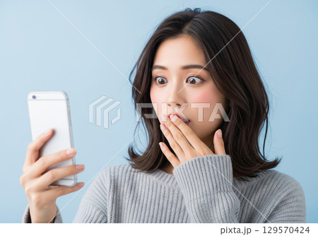 スマートフォンを見て驚き、片手で口をおさえる女性の表情 129570424