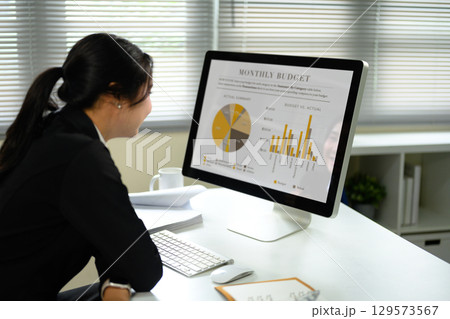 Female office worker reviewing a monthly budget report with pie and bar charts on a desktop screen 129573567