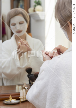 鏡の前でフェイスマスクで肌ケアしながらネイルを塗るシニア女性 129574601
