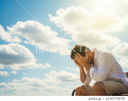 Man hides face in hands sitting outdoors in sweltering heatwave, sun shining hard above with little shade or relief 129574976