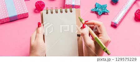 Top view of female hand making some notes in noteebok on pink background. New Year decorations and toys. Christmas time concept 129575229