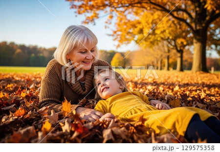 Grandmother and granddaughter lying joyfully in fallen golden leaves enjoying a peaceful autumn afternoon together outdoors 129575358