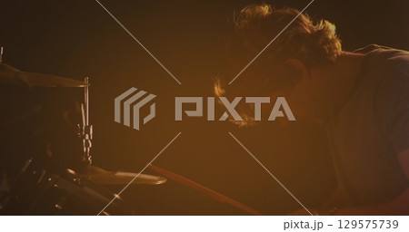 Image of caucasian male drummer playing drum kit, under orange coloured lights 129575739
