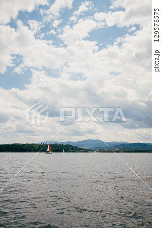 Sailboats with red and white sails are gracefully navigating serene lake, tranquil water reflecting cloudy sky above and distant mountains adding to picturesque scenery. Sailboats gliding across lake 129578575