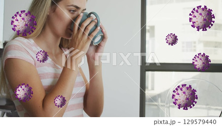Image of coronavirus cells over caucasian young woman drinking coffee while working at desk 129579440