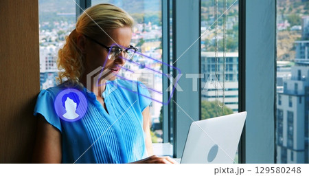 woman in blouse and glasses typing on laptop near office window, with holographic icons, copy space 129580248