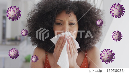 Image of coronavirus cells rotating over woman with afro hair sneezing with tissue 129580284