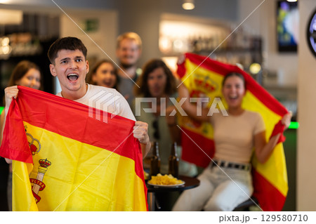Excited spanish fans cheer for their team as they watch the match on TV in beer bar 129580710