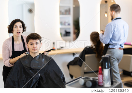 Adult woman hairdresser gives haircut to young guy 129580840