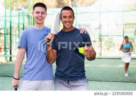 Portrait of satisfied two man with racket and ball in his hands on padel court Portrait of satisfied two man with racket and ball in his hands on padel court 129580988