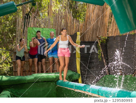 Cheerful girl crossing pool of water on inflatable log with hanging obstacles Cheerful girl crossing pool of water on inflatable log with hanging obstacles 129580993