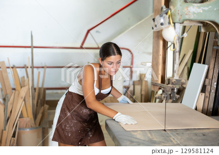 Girl employee of workshop cuts wooden board into parts using saw 129581124