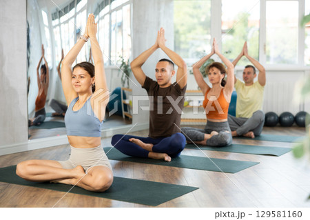 Woman doing lotus pose of yoga on black mat in gym room with other persons 129581160