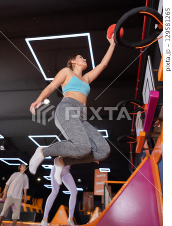 Happy young female in blue top and gray leggings, throwing ball in basketball hoop while jumping on trampolines in entertainment center 129581265