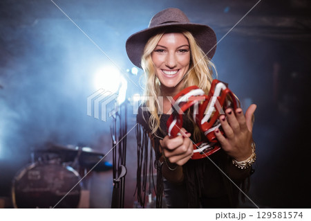 Female musician standing on stage holding red-and-white tambourine with stage lights glowing behind 129581574