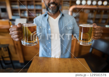 Mature Indian man wearing denim shirt holding two frothy beer mugs at wooden bar counter 129581578