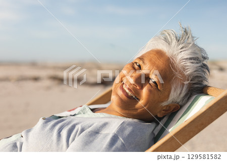 Senior woman reclining on wooden lounge chair with striped cushion on beach in sun, copy space Senior woman reclining on wooden lounge chair with striped cushion on beach in sun, copy space 129581582