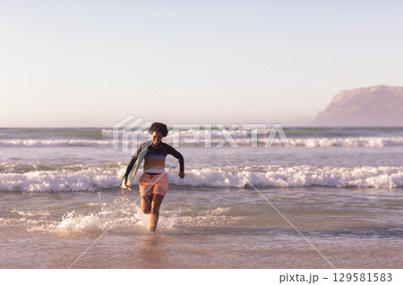 African American woman running from surf toward sandy shore holding surfboard wearing rash guard African American woman running from surf toward sandy shore holding surfboard wearing rash guard 129581583