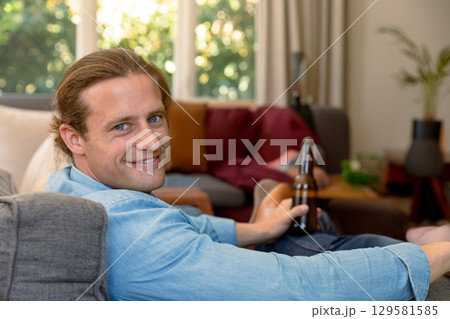 Male reclining on gray sofa in living room with potted plant, holding dark beer bottle 129581585