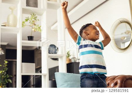 African American boy celebrating with raised fists standing on sofa in living room, copy space 129581591