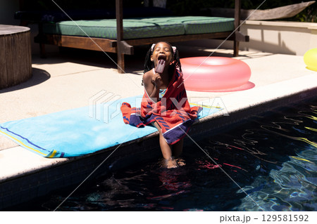African American girl sitting wrapped in red towel at pool edge licking popsicle under sunlight 129581592