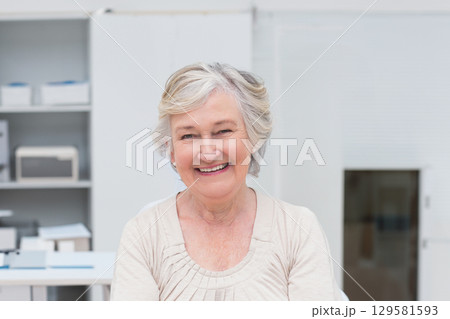 Smiling senior female sitting at desk in home office sorting paperwork near printer and shelving 129581593