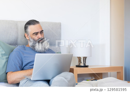 Senior man leaning against tufted headboard and working on laptop on bed, copy space 129581595
