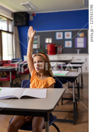 Elementary school girl raising her hand at desk in classroom with pencil and open notebook 129581596