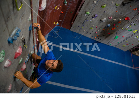 Male climber wearing blue shirt ascending gym wall with multicolored holds, safety rope, copy space 129581613