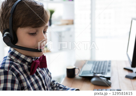 School-age boy wearing red bow tie and headset typing on keyboard in home office, copy space 129581615