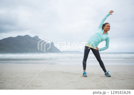 Woman performing side bend at sandy beach waterline in sports jacket and running shoes, copy space Woman performing side bend at sandy beach waterline in sports jacket and running shoes, copy space 129581626