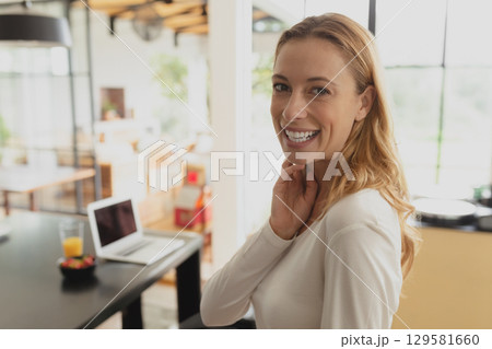 Woman standing beside dining table under pendant lamp in open-plan home with laptop, juice, fruit Woman standing beside dining table under pendant lamp in open-plan home with laptop, juice, fruit 129581660