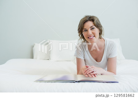 Mid adult woman lying on white linens with pillows in bedroom reading book and wearing t-shirt Mid adult woman lying on white linens with pillows in bedroom reading book and wearing t-shirt 129581662