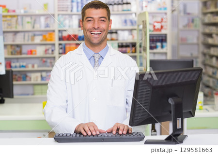 Male pharmacist standing at pharmacy counter typing on keyboard with shelves of medication Male pharmacist standing at pharmacy counter typing on keyboard with shelves of medication 129581663