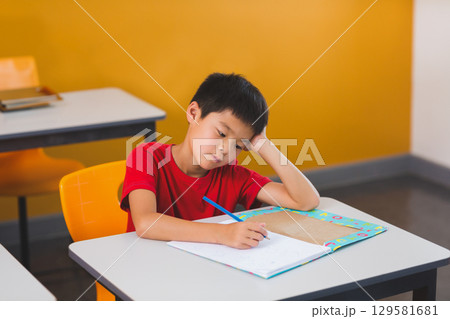 Asian boy resting head on hand writing in colorful workbook at school desk with pencil Asian boy resting head on hand writing in colorful workbook at school desk with pencil 129581681