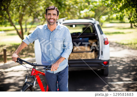 Man standing holding red bicycle next to white SUV with open hatch in sunlit park lot Man standing holding red bicycle next to white SUV with open hatch in sunlit park lot 129581683