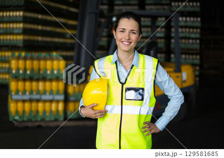 Female warehouse supervisor standing in storage wearing safety vest holding hardhat with forklift 129581685