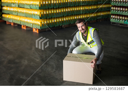 Male warehouse worker wearing vest kneeling in warehouse lifting box beside crates, copy space 129581687