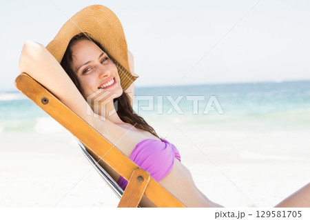 Woman reclining on lounge chair at beach wearing purple bikini top and straw hat, copy space Woman reclining on lounge chair at beach wearing purple bikini top and straw hat, copy space 129581705