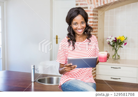 African American woman sitting on wooden countertop in kitchen holding tablet and pink coffee mug African American woman sitting on wooden countertop in kitchen holding tablet and pink coffee mug 129581708