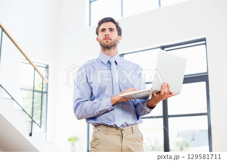 Man holding open laptop and standing in modern office space by staircase under bright windows Man holding open laptop and standing in modern office space by staircase under bright windows 129581711