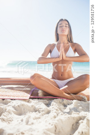 Woman wearing white swimsuit practicing yoga sitting cross-legged on bright mat on beach with waves 129581716