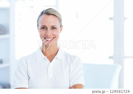 Mid adult woman standing with arms crossed smiling in bright office wearing white polo shirt 129581724